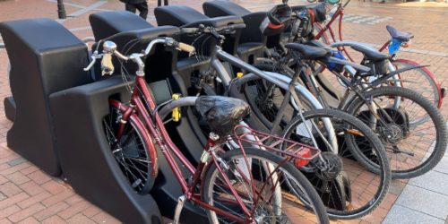 Several bicycles of various styles and colours parked in a modern black moulded bike stand on a brick-paved urban street.