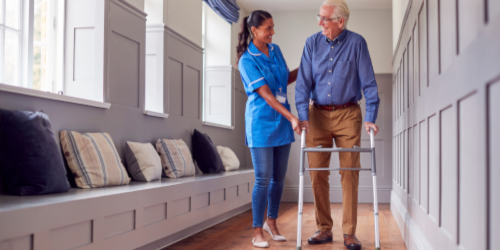 A healthcare worker in blue scrubs assists an elderly man using a walking frame along a bright hallway, with a built-in bench seat and decorative cushions along the wall.