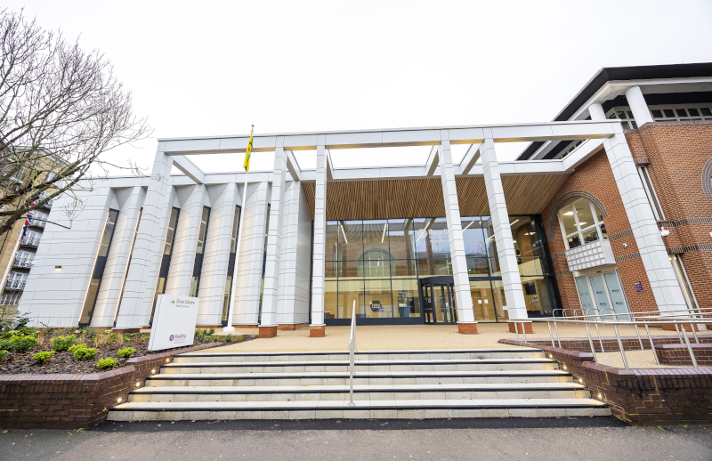 Exterior of a modern civic building with white columns, glass entrance doors, and a yellow flag, approached by wide stone steps with brick detailing on the right wing.