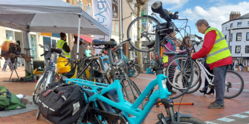 A Dr Bike community cycle repair event in a town square, with several bicycles including a turquoise cargo bike in the foreground. Volunteers in hi-vis vests inspect and repair bikes beneath a white gazebo, with red-brick buildings visible in the background.