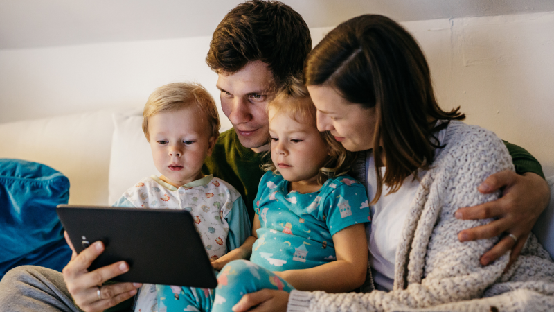 A family of four — two parents and two young children in pyjamas — sit closely together on a sofa, looking at a tablet screen.