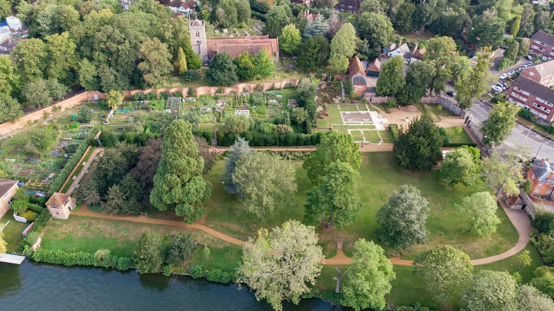 Aerial view of Caversham Court Gardens beside the River Thames, showing a walled garden with vegetable plots, a lawn with mature trees and winding paths, a small stable building, and a church with a tower among surrounding trees.