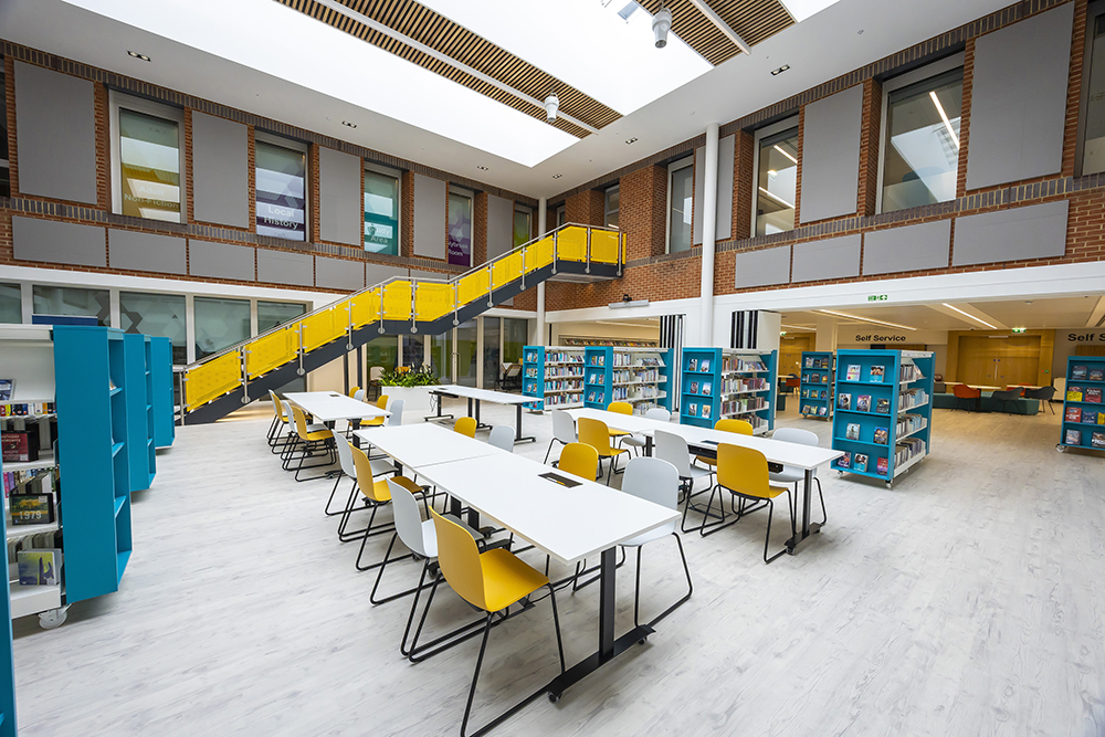 Open-plan Central Library interior with study tables, bookshelves, and a striking yellow staircase beneath a large skylight.