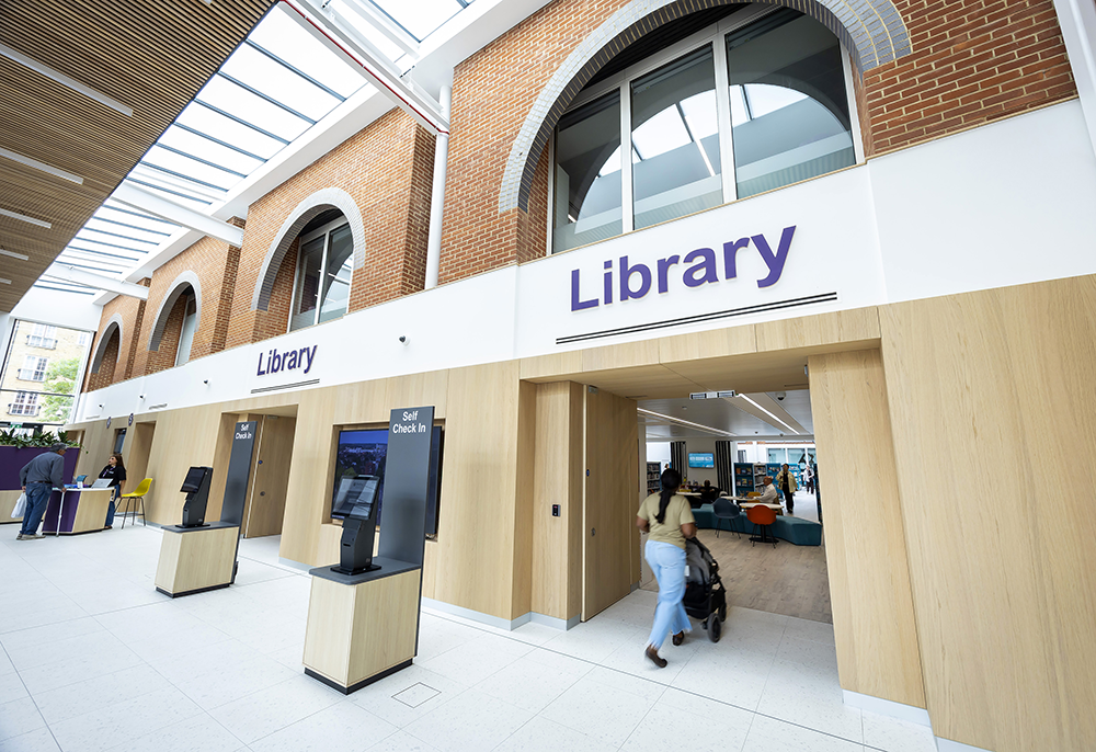 Entrance to the Central Library with large arched brick windows, self-service check-in kiosks, and a visitor entering the bright, modern library space.
