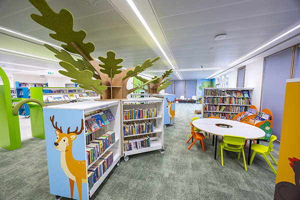 Children's library with colourful woodland-themed bookshelves, low reading tables and chairs, and a bright, welcoming space for young readers.