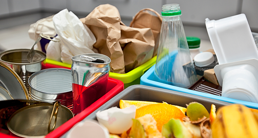 A close-up of household recycling sorted into colorful plastic bins, including empty cans, a plastic bottle, paper bags, cardboard, and food scraps.