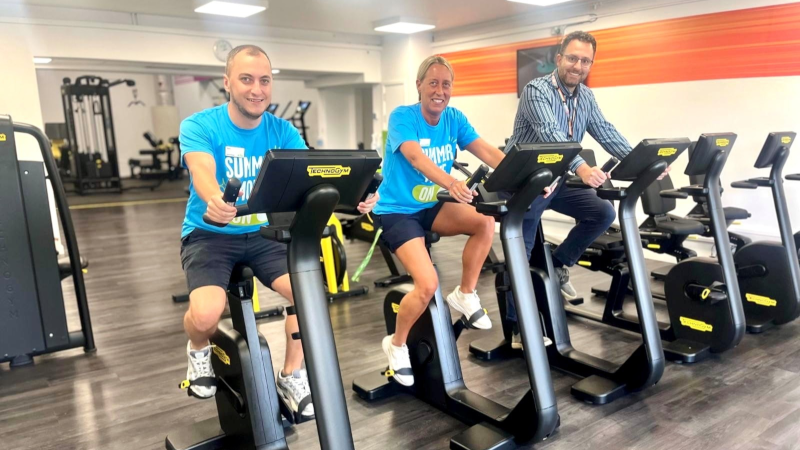 Three people smiling while riding exercise bikes in a gym, two wearing blue 'Summer' campaign T-shirts, the other in shirt with lanyard - this one is Councillor Jacopo Lanzoni. Rows of gym equipment are in the background.