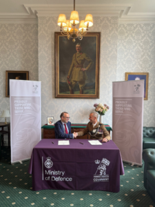 Two people shake hands at an Armed Forces Covenant signing ceremony, seated behind a Ministry of Defence branded table.