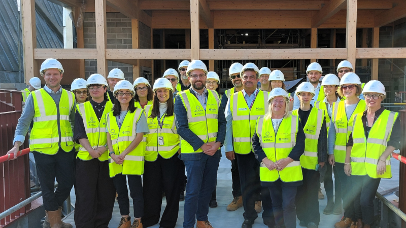 Group photo of around 20 people wearing high-visibility yellow safety vests, white hard hats and protective footwear, standing on a newly completed pedestrian walkway at a construction site. The group faces the camera in front of the timber-framed structure of the new Studio Theatre building. Red railings line the walkway on either side, and the partially completed theatre can be seen in the background with exposed wooden beams, brickwork and construction materials visible behind the group. Sunlight illuminates the scene, highlighting the progress of the development.