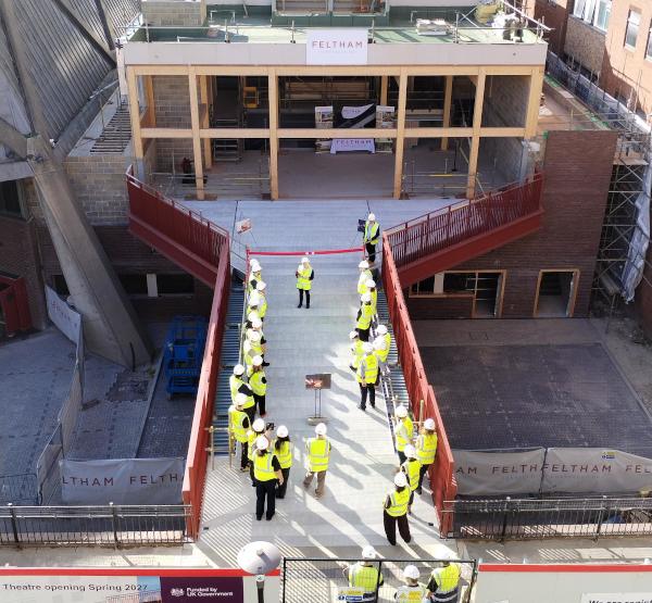 Aerial view of a newly constructed pedestrian bridge leading to a building under construction. Around 20 people wearing high-visibility jackets and hard hats stand along both sides of the bridge and at its entrance, appearing to participate in an opening or inspection event. The bridge has red railings and connects to a partially completed building displaying the name “Feltham.” Construction materials and scaffolding are visible around the site, and a sign at the bottom of the image reads “Theatre opening Spring 2027.”