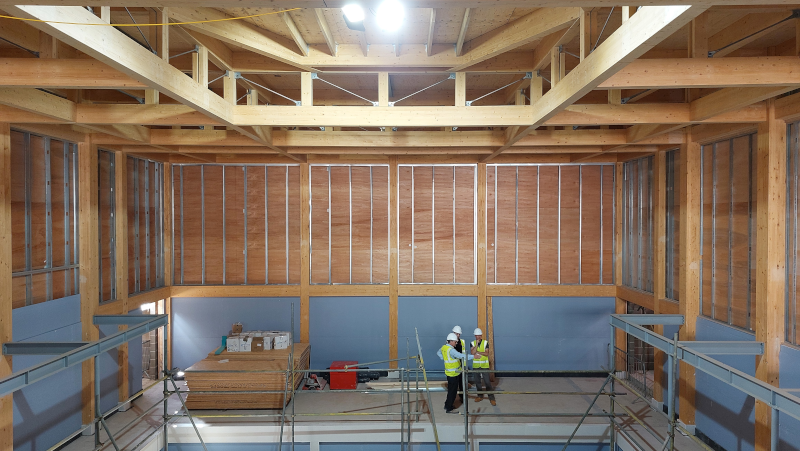 Interior view of the new Studio Theatre under construction, showing the auditorium space from an upper level. The building features a large exposed timber frame with substantial wooden beams, columns and ceiling supports. Scaffolding and temporary safety barriers line the lower level, where construction work is ongoing. Stacks of building materials and equipment are visible on the left side of the space, while two people wearing high-visibility safety vests and white hard hats stand on the floor below. The layout shows the developing theatre structure, including the gallery level overlooking the main performance area. Bright overhead lighting and natural light highlight the scale and progress of the construction.