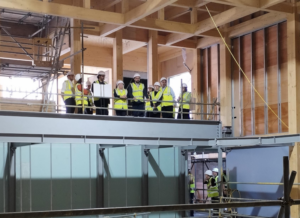 Interior view of the Studio Theatre construction site. A group of people wearing high-visibility safety vests and white hard hats stand on an upper-level platform behind temporary safety barriers during a site visit. The building’s timber frame is clearly visible, with large wooden beams and columns supporting the structure. Scaffolding is positioned on the left side of the image, while exposed wall panels, steel supports and unfinished building services can be seen throughout the space. Additional visitors in safety equipment are visible at ground level below. Natural light enters through large openings in the building, highlighting the progress of the auditorium and gallery areas as construction continues.