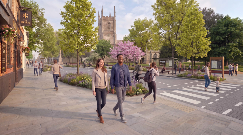 A landscaped town-centre street beside a public square and church, featuring wide stone pavements, mature trees, flower beds and a zebra crossing. Several pedestrians are walking through the area, while a cyclist travels along the road. A historic church tower rises in the background behind green space and pink-blossoming trees, creating an attractive and welcoming urban setting.