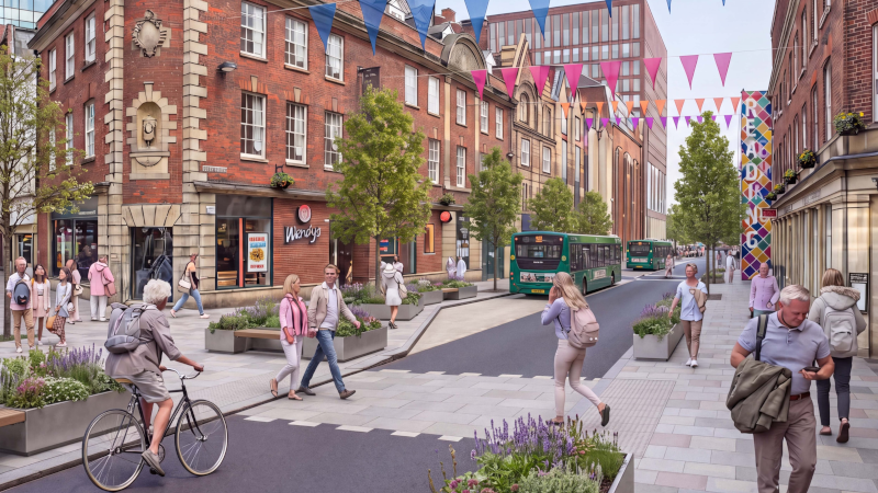 A pedestrian-friendly city street lined with red-brick buildings, shops and cafés, featuring wide pavements, planted seating areas and colourful bunting strung overhead. People walk, cycle and cross the street while two green buses travel along a single carriageway. Trees, flower planters and a brightly patterned building facade contribute to a vibrant, welcoming urban environment.