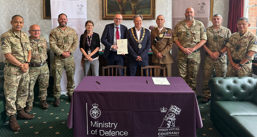 Group photograph of military personnel and civic representatives at an Armed Forces Covenant signing ceremony, with a Ministry of Defence table and covenant banners in the background.